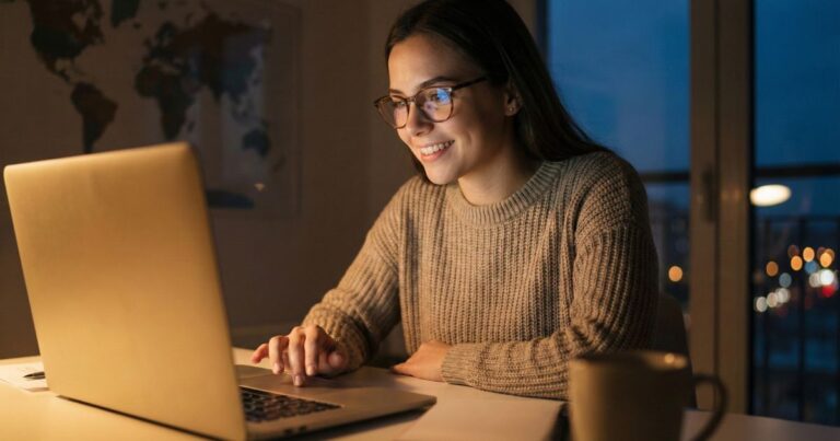 Young blogger publishing first blog post at night, laptop screen glowing warmly as they smile with excitement about reaching a global audience and building a successful blog.