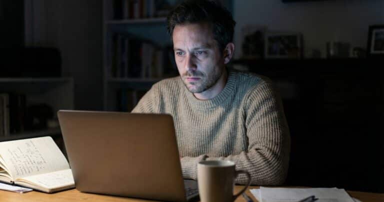 Course creator staring at laptop in dim home office after AI rewrite, open notebook on desk at night